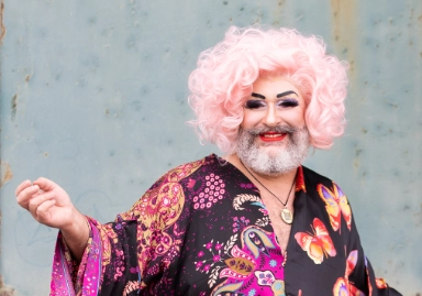 This image shows a person with a vibrant and playful appearance, featuring a pink curly wig, dramatic makeup, and a beard. They are wearing a colorful robe with butterfly and paisley patterns, smiling confidently against a weathered blue background.