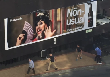 This image shows a billboard advertisement for '@Hoptsoda,' featuring a person in dramatic drag makeup, a tiara, and holding a pink fan. The text on the billboard reads 'Non-usual' and 'Lower-sugar soda,' with an image of the product on the right. Pedestrians are seen walking below the ad on the street.