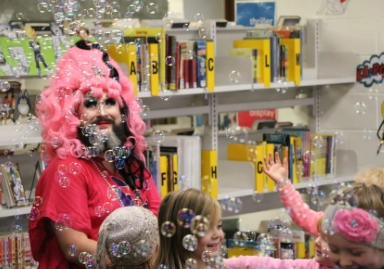 This image features a lively scene in a library, where a person with a bright pink wig, dramatic makeup, and a beard interacts with children surrounded by floating bubbles. Shelves filled with books labeled with letters are visible in the background, adding to the vibrant, playful atmosphere.