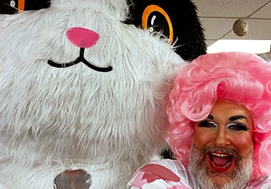 A person with a glittery beard, bright pink wig, and dramatic makeup poses with a wide, joyful smile next to a large, plush mascot resembling a black-and-white cat with big orange eyes. The person is wearing a white and pink outfit.